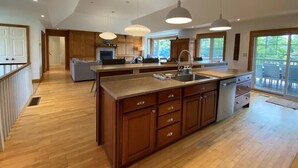 A spacious kitchen with wooden cabinets, a large island with a sink, pendant lights, and a view of the outdoors through large windows. - Private Beach, Hot Tub, A Vacay to Remember! (Prince Edward)