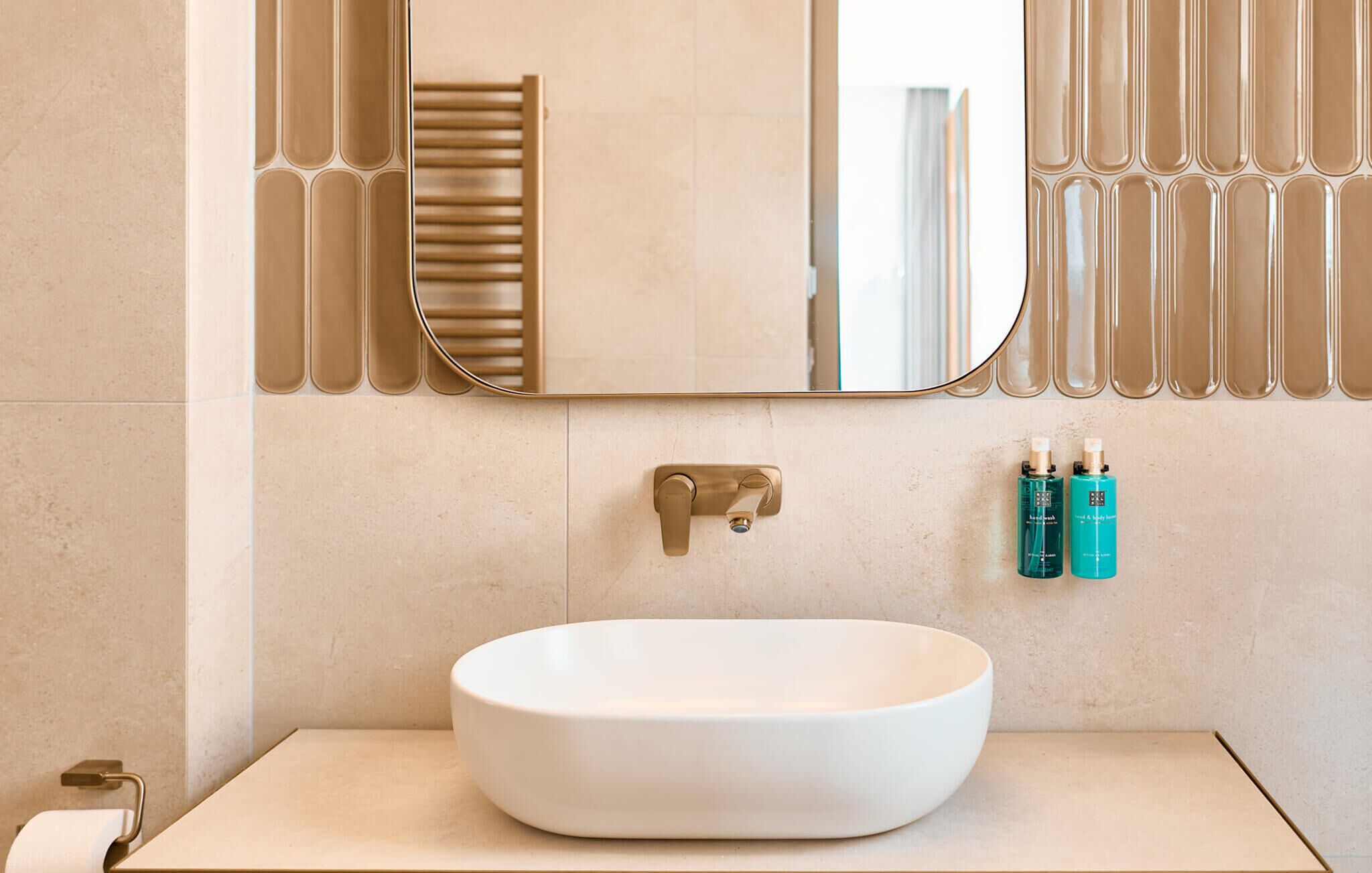 Ein Badezimmer mit einem weißen Waschbecken, einem goldenen Wasserhahn, einem Spiegel mit dekorativem Rahmen und zwei Shampooflaschen an der Wand.