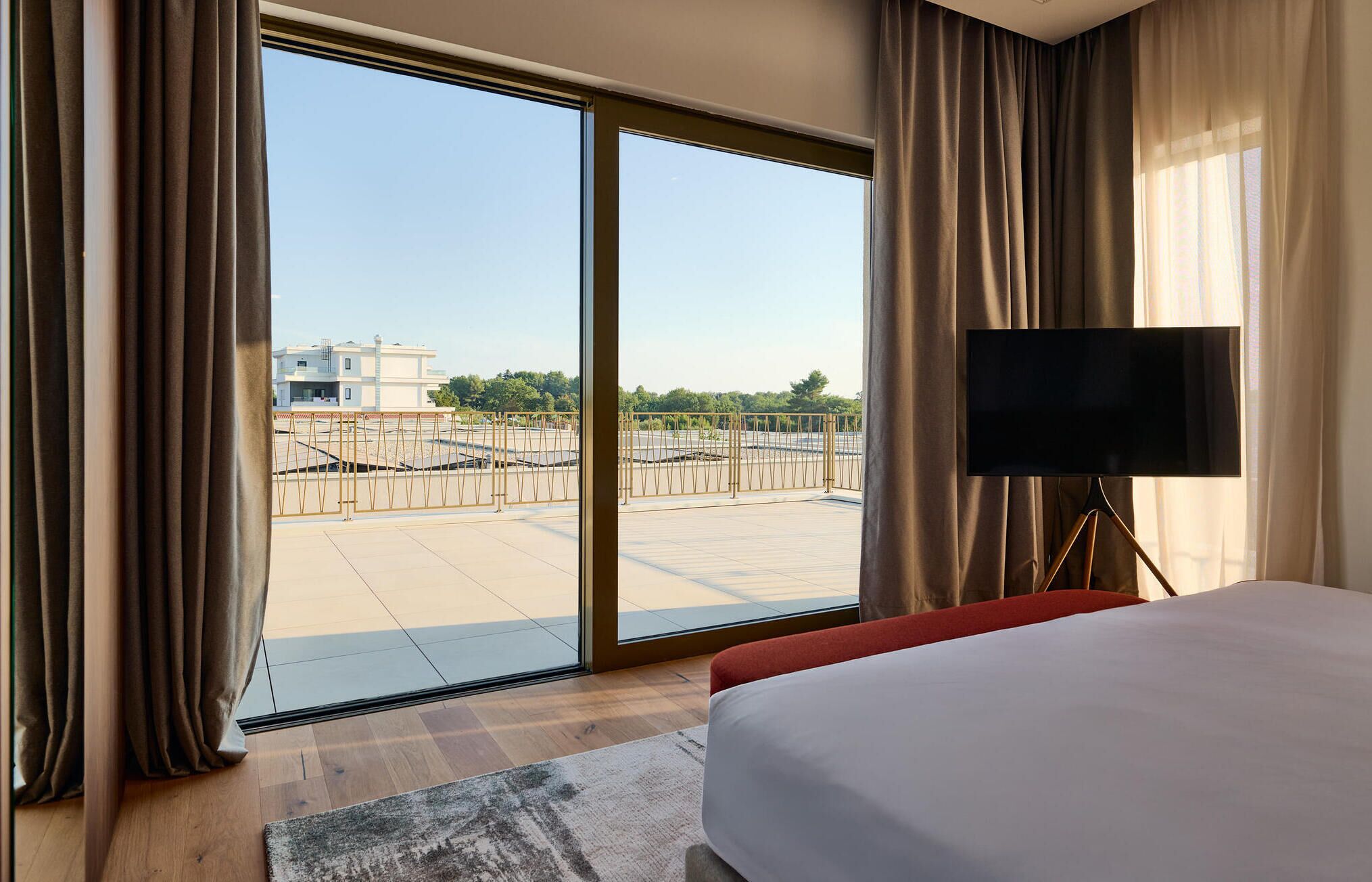 A hotel room with a large sliding glass door leading to an outdoor terrace, a bed, a TV on a stand, and a view of a building and trees.