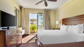 A hotel room with a bed, a TV, a desk with a lamp, and a view of palm trees through the door. - Tropical 2BR Partial Ocean View Villa at The Point at Poipu (Koloa)