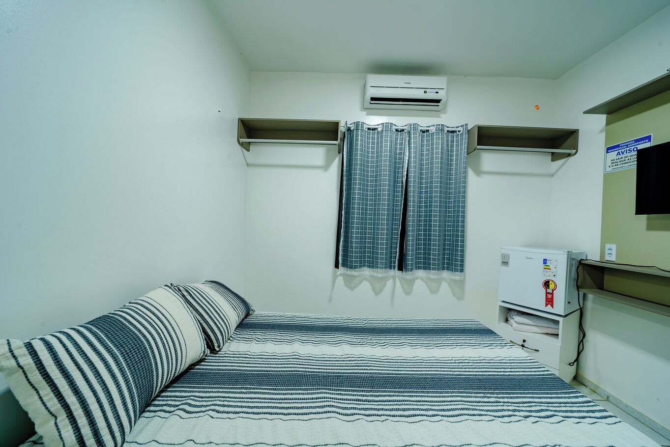 A small, sparsely furnished room with a bed, a pillow, a wall-mounted air conditioner, a television, and a small cabinet.