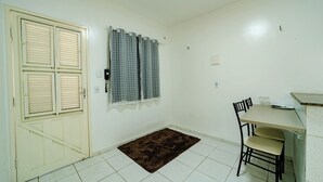 Una habitación con puerta, ventana con cortinas, suelo de baldosas, alfombra marrón, una mesa con sillas y una pequeña cocina.
