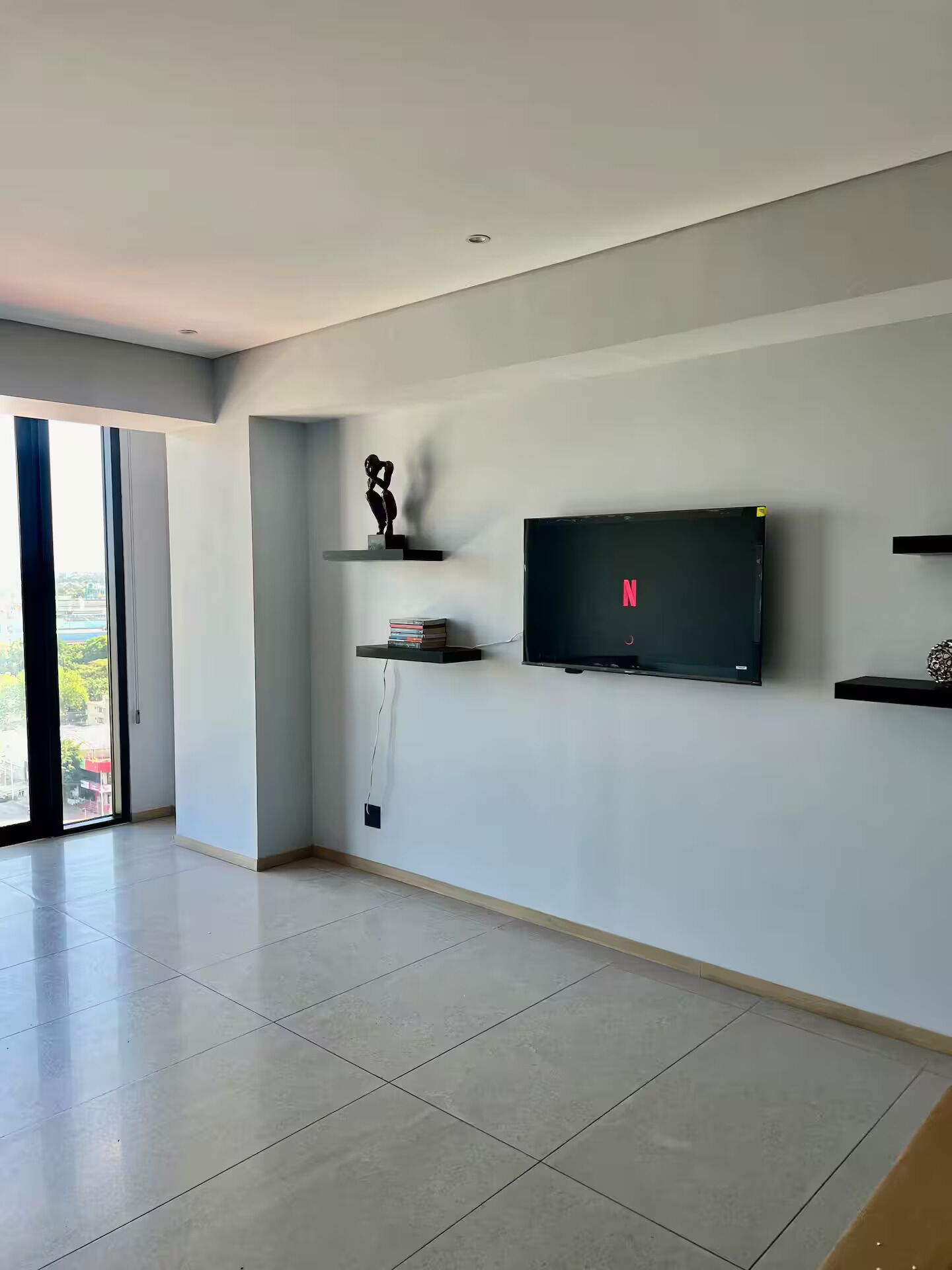 Una sala de estar moderna con un televisor de pantalla plana montado en la pared, una escultura en un estante y vistas a través de un gran ventanal corredero.