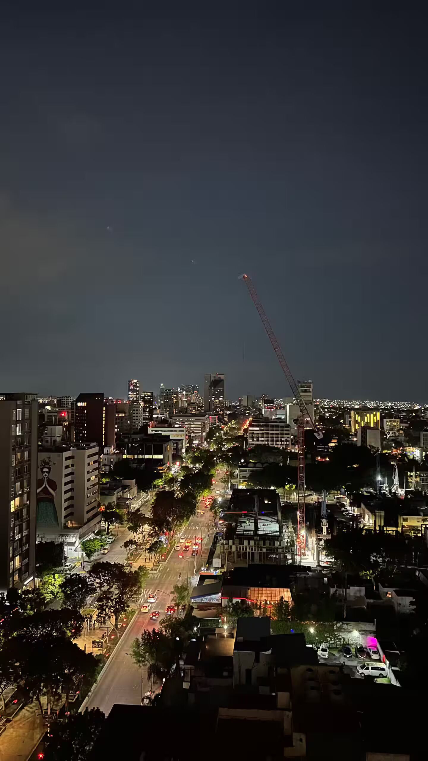 A nighttime cityscape with illuminated buildings, a prominent crane, and a busy street.