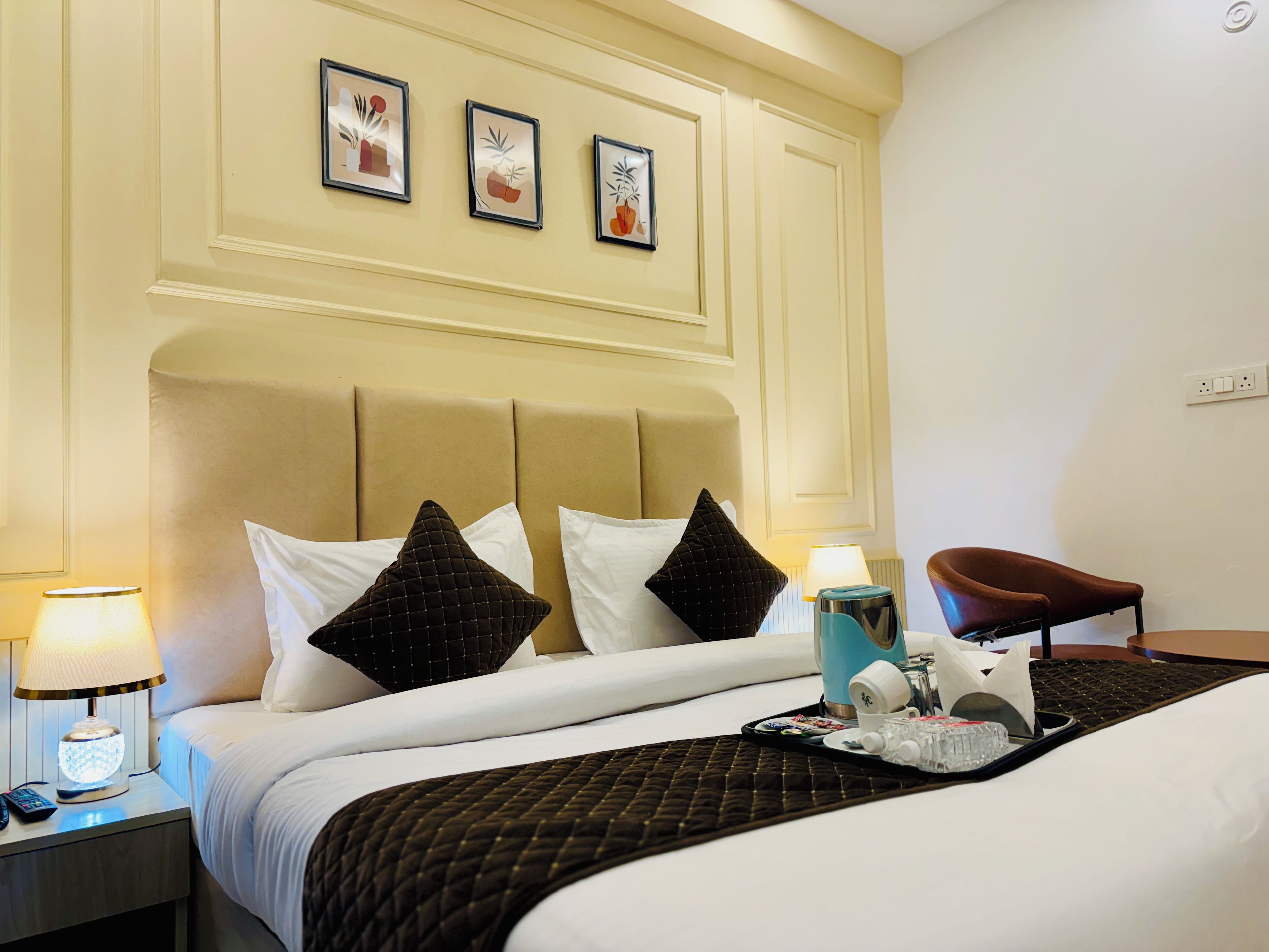 A hotel room with a bed, nightstand, lamp, chair, and wall-mounted artwork.
