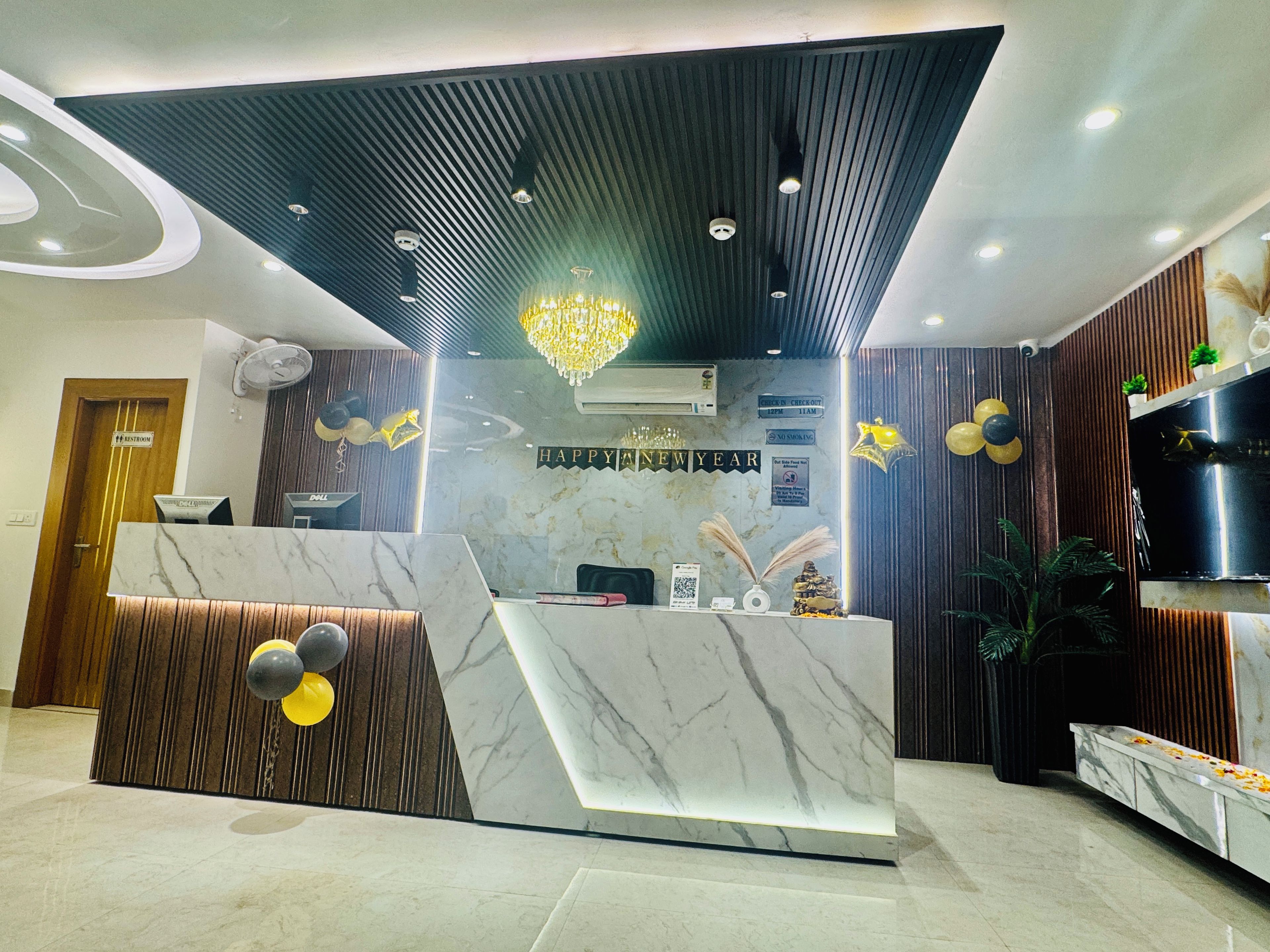 A reception desk with a marble top, a wooden panel design, and a chandelier. There is a sign that reads "HAPPY NEW YEAR" and a "CHECK IN/ CHECK OUT" sign.