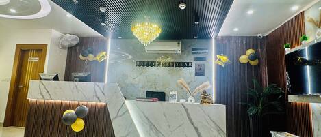 A reception desk with a marble top, a wooden panel design, and a chandelier. There is a sign that reads "HAPPY NEW YEAR" and a "CHECK IN/ CHECK OUT" sign.