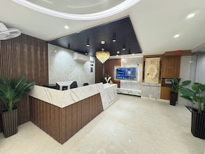 A reception area with a marble counter, wooden paneling, a mounted TV, and a chandelier. - Hotel Classic Crowne (New Delhi)