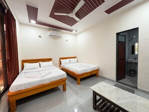 A hotel room with two wooden beds, a ceiling fan, an air conditioning unit, a small table, and a bathroom with a mirror and sink. - Sandalwoods Resorts (Karjat)