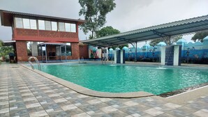 A swimming pool with a tiled deck, a covered area with a roof, and a building with large windows in the background.