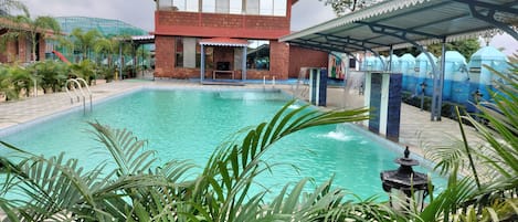 Una piscina con zona cubierta, rodeada de palmeras y un edificio de techo rojo.