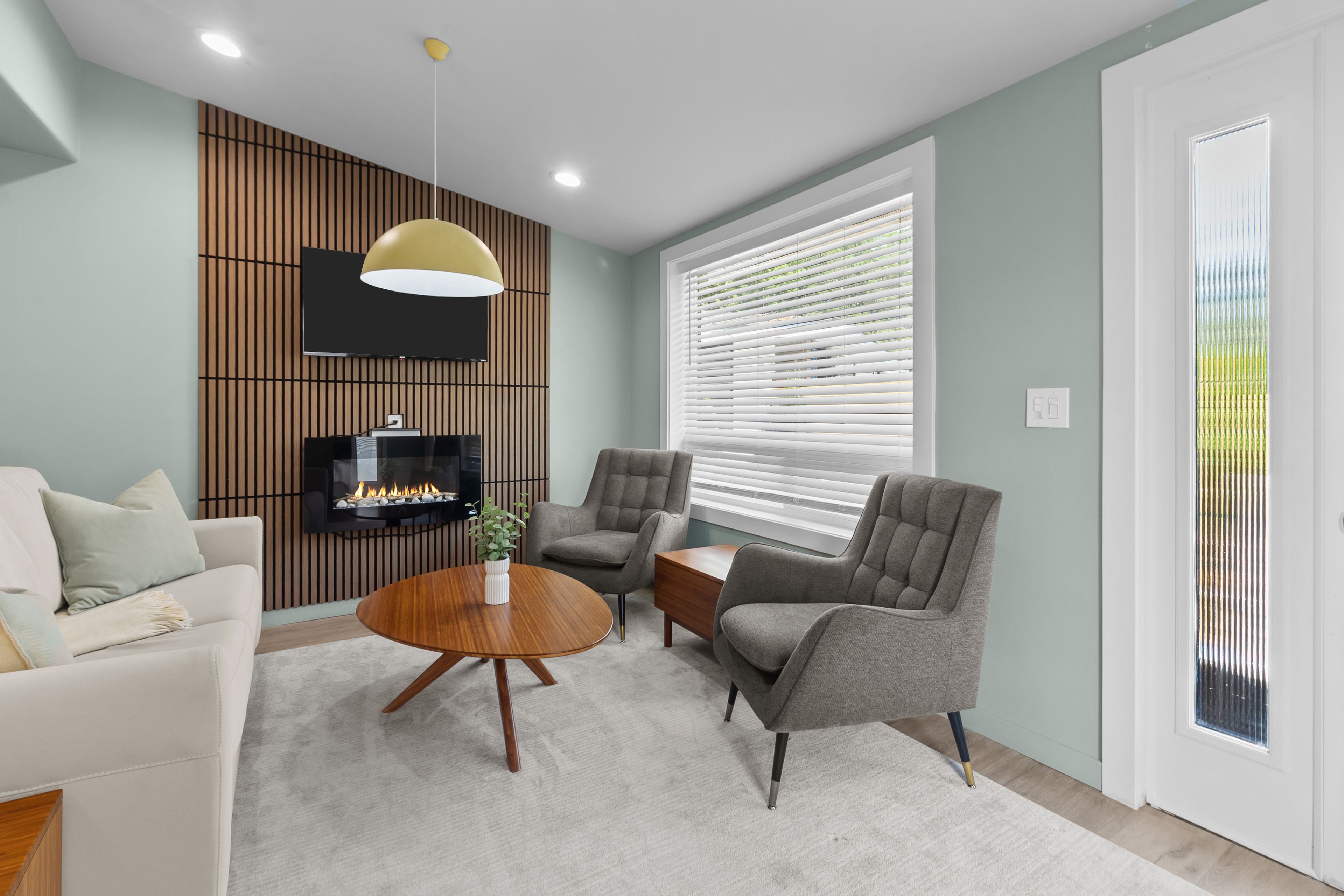 A modern living room with a flat-screen TV, a fireplace, grey armchairs, a white sofa, and a large window with blinds.