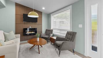 A modern living room with a flat-screen TV, a fireplace, grey armchairs, a white sofa, and a large window with blinds.
