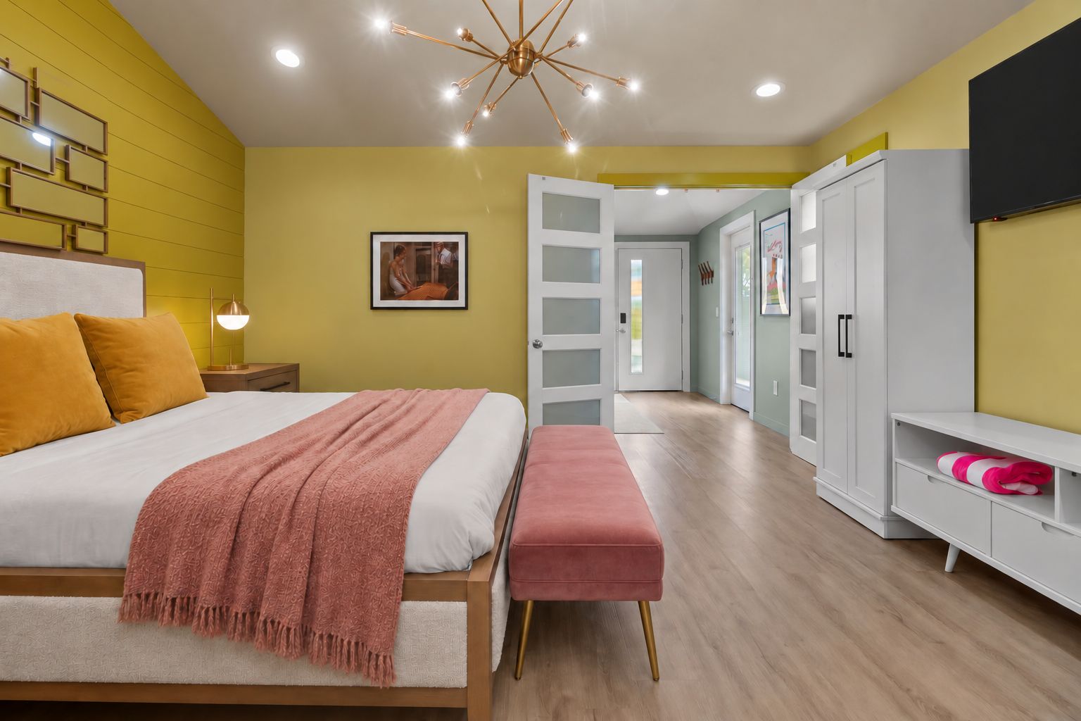 Une chambre moderne avec un lit, une table de chevet et un banc. On y trouve une armoire avec un miroir et une étagère sur laquelle est posée une serviette rose.