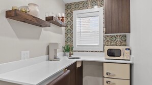 A compact kitchen with a white countertop, a microwave, a small refrigerator, and a window with blinds.