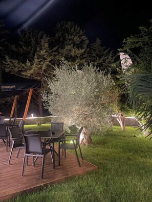 An outdoor dining area with tables and chairs, a small tree, and a stone wall in the background.