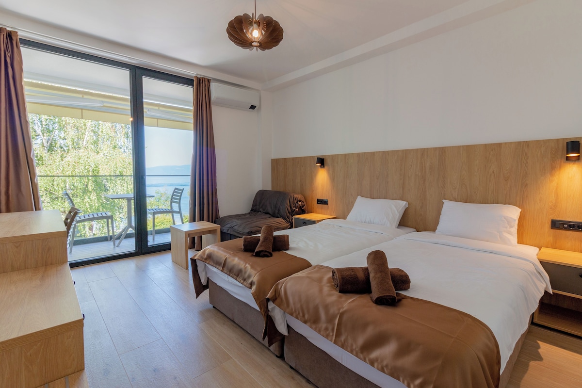 A modern hotel room with a large bed, a desk, and a balcony with chairs.