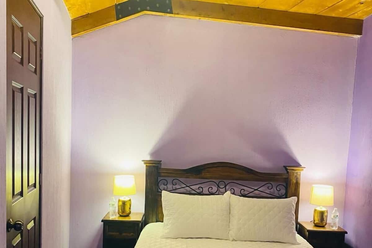 Ein Einzelbett mit gesteppter Bettdecke, zwei Nachttischlampen, ein Holz-Kopfteil und eine Tür links.