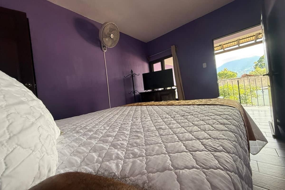 Ein Schlafzimmer mit einem großen Bett, einem Standventilator und Blick auf einen Balkon mit Geländer.