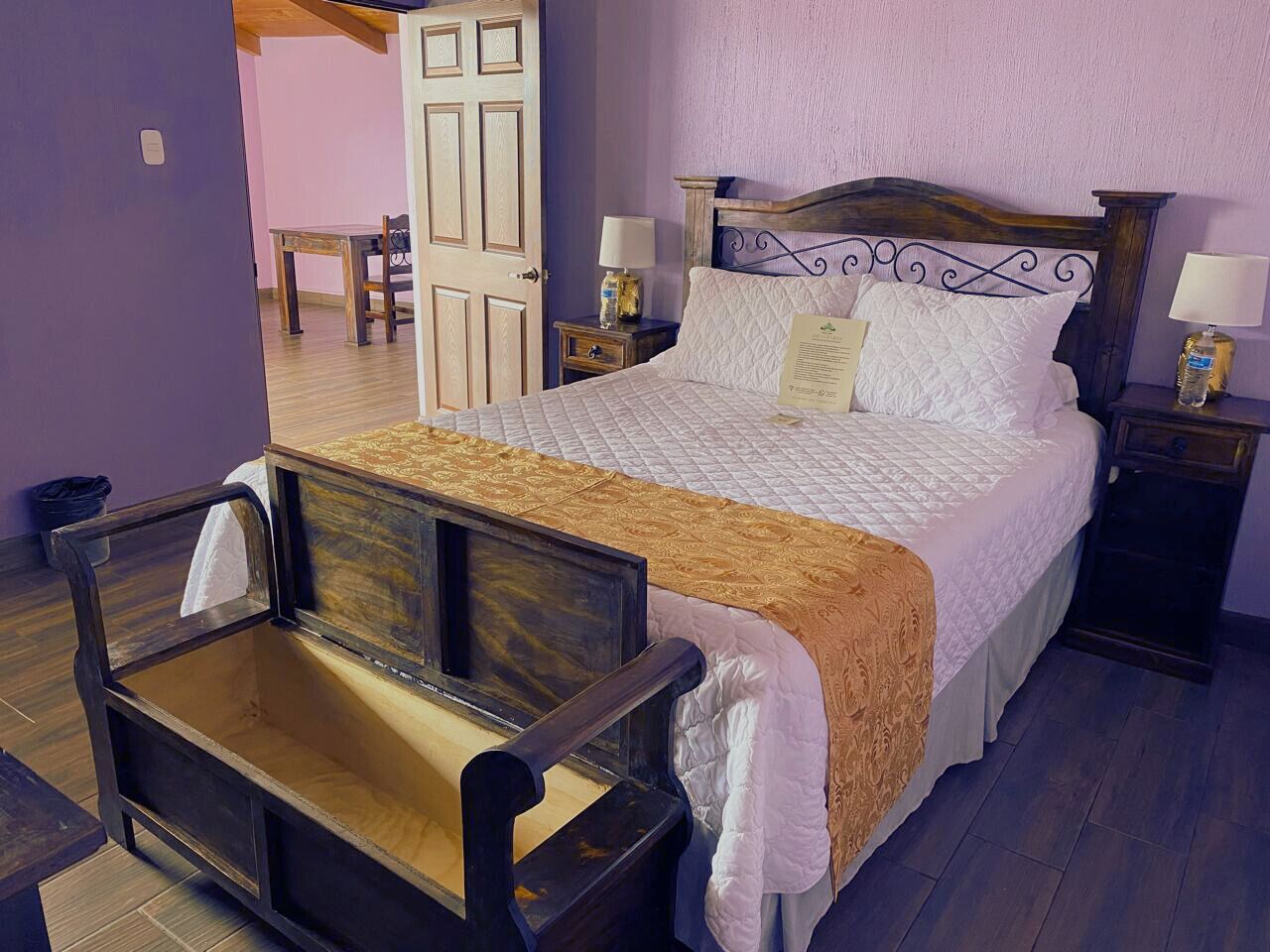 Un dormitorio con cama, mesitas de noche, un banco de madera y una puerta que da a otra habitación.