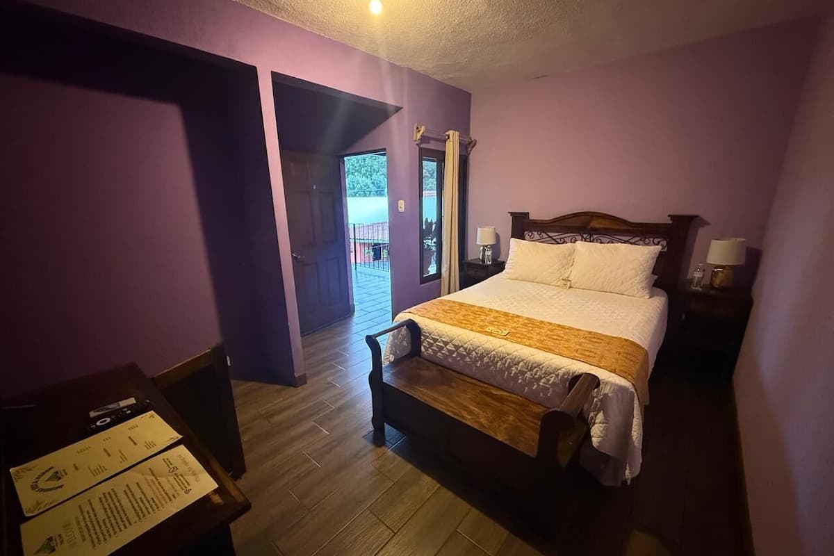 Ein Hotelzimmer mit einem Bett, einem Schreibtisch und einem Sessel. Ein Fenster mit Vorhängen und eine Tür zu einem Außenbereich.