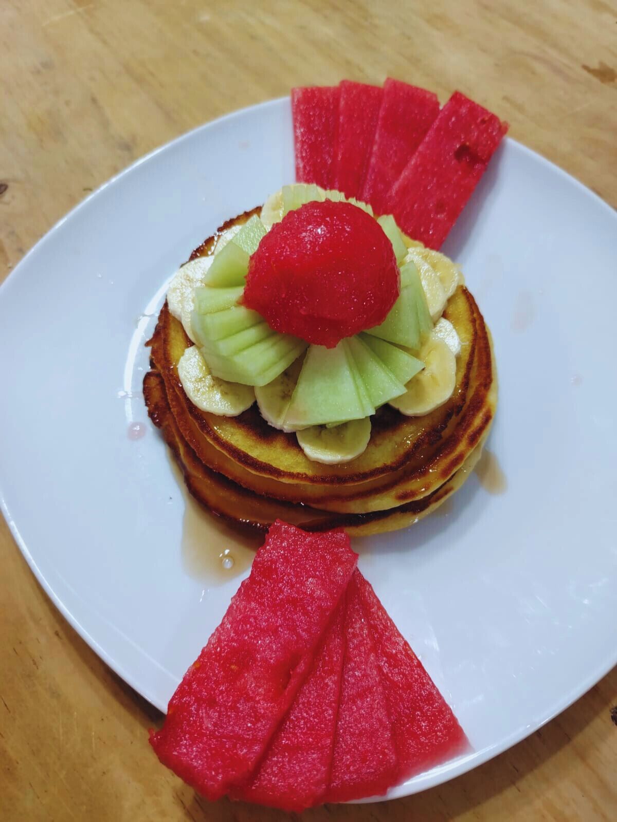 A plate with pancakes topped with sliced bananas, a strawberry, and watermelon slices.