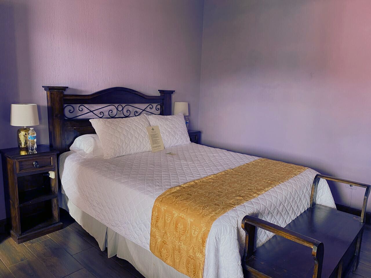 Una cama bien hecha con colcha blanca y manta dorada, flanqueada por mesitas de noche de madera oscura con lámparas y una botella de agua.