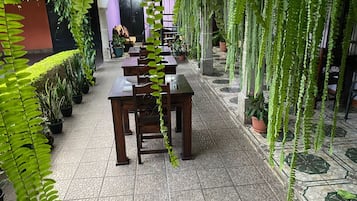 A covered outdoor seating area with tables and chairs, surrounded by potted plants and hanging greenery.
