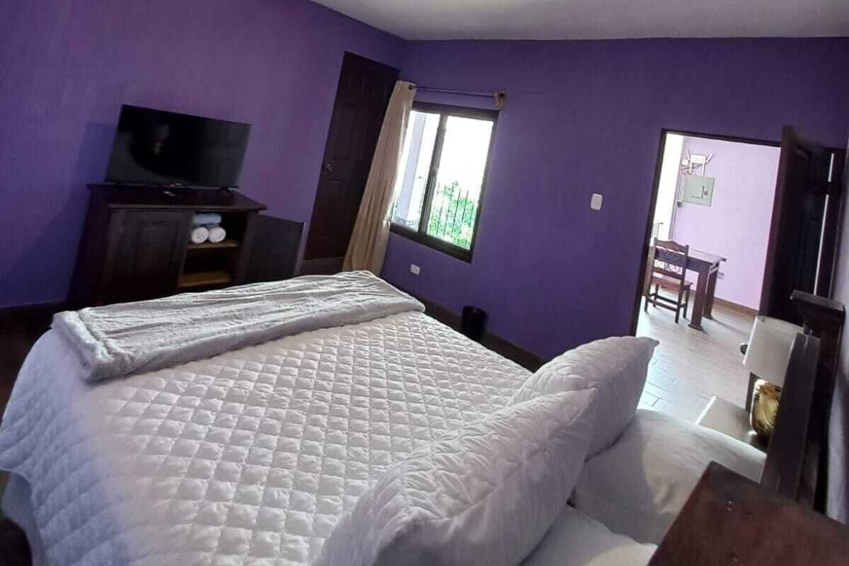 Ein Zimmer mit einem Bett, einem Fernseher auf einem Schrank, einem Fenster mit Vorhängen und einer Tür zu einem weiteren Raum.