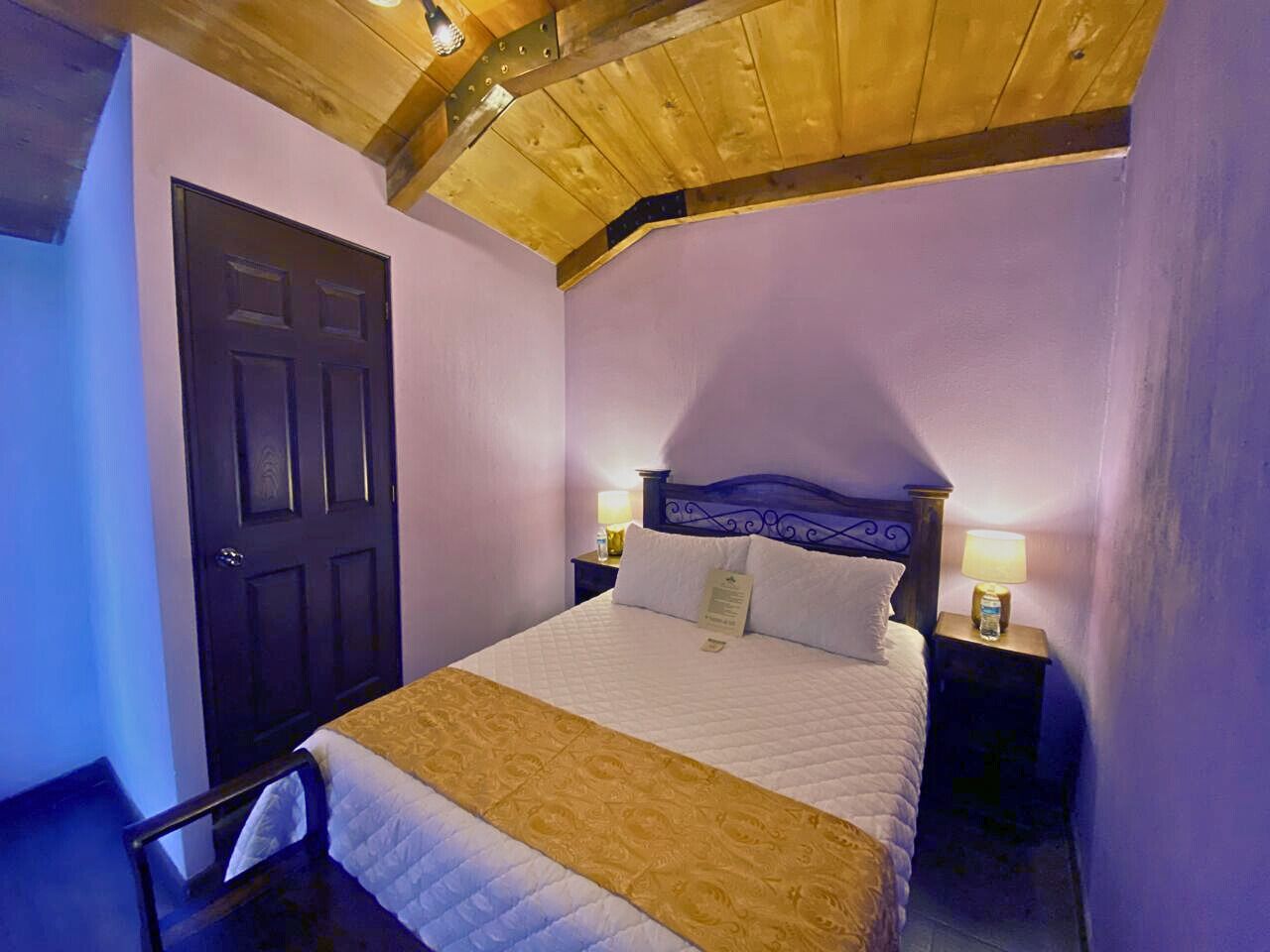 Une chambre avec un plafond en bois, un lit recouvert d’une couvre-lit blanche et dorée, deux tables de chevet avec des lampes et une porte sombre.