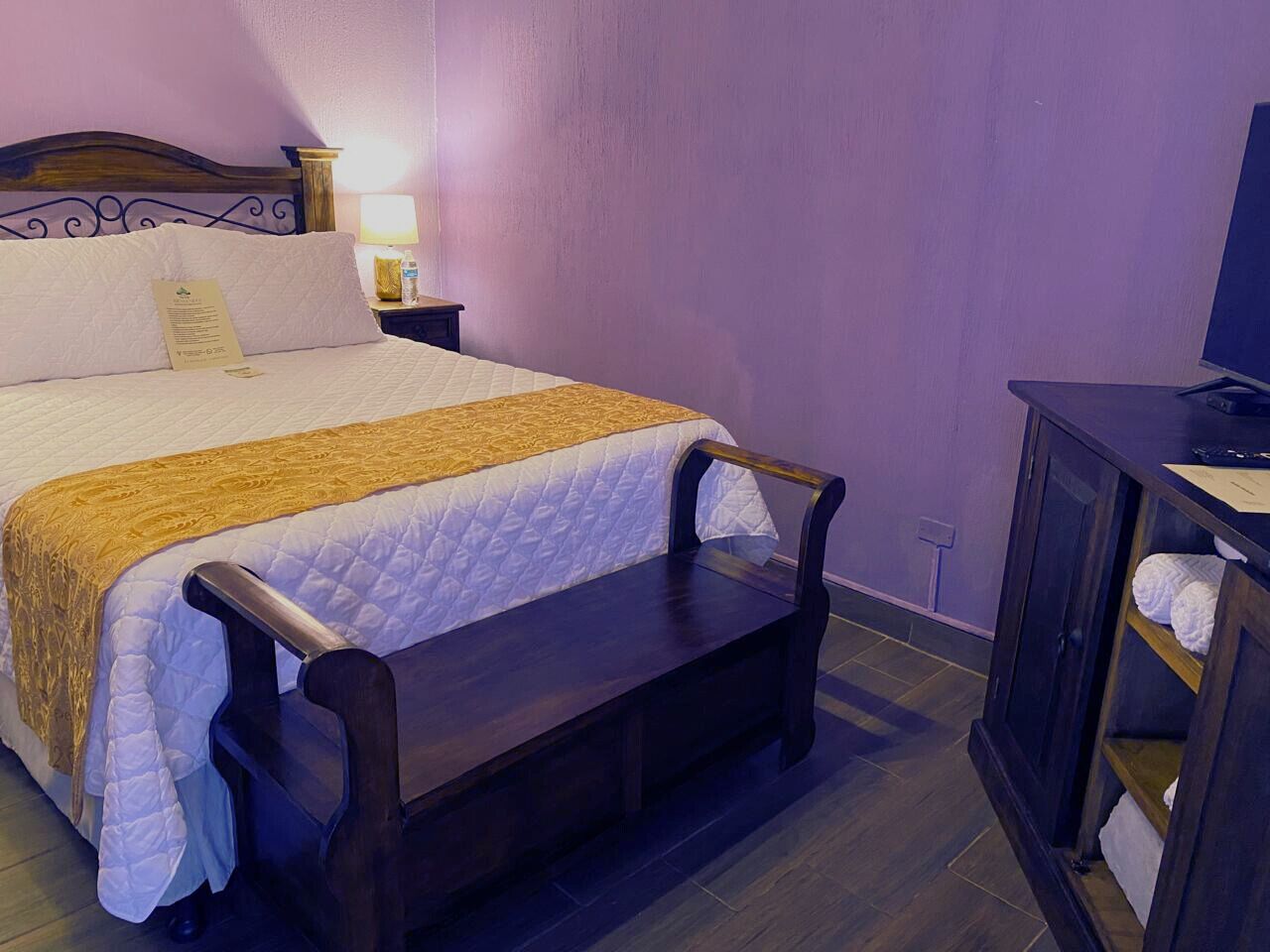 A neatly made bed with a wooden headboard, a bedside table with a lamp, and a bench at the foot of the bed.