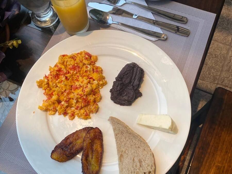 Een ontbijtbord met roerei, zwarte bonen, gebakken plantanen en toast, vergezeld van een kop koffie en een glas sinaasappelsap.