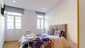 Een slaapkamer met een groot bed, twee ramen, een radiator en een aan de muur bevestigde televisie.