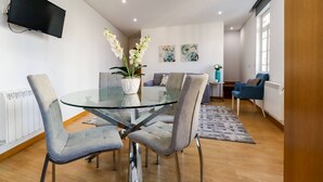 Een moderne woonkamer met een eettafel met glazen blad, omringd door grijze fauteuils, een flatscreen televisie aan de muur en twee abstracte schilderijen.