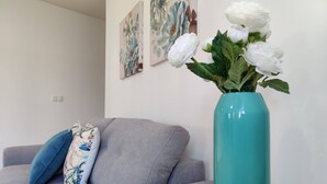 Een woonkamer met een grijze bank, een turquoise vaas met witte bloemen, en een klein wit tafeltje met een kaars en een decoratief vliegtuigbeeldje.