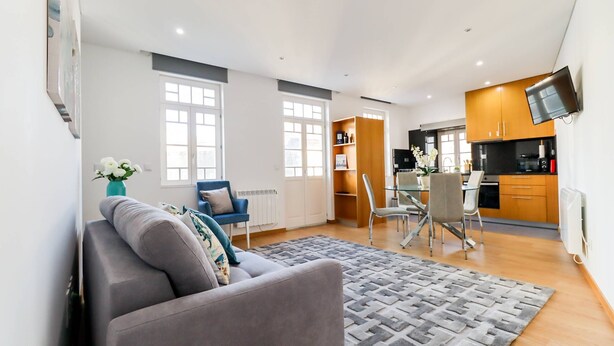 Een moderne woonkamer met een grijze bank, een kamerkleed met patroon en een eethoek met een glazen tafel en stoelen.