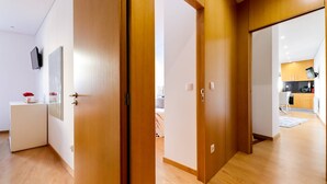 Een modern appartement met houten vloeren, een witte, aan de muur bevestigde televisie, een klein wit kastje, en een keukenruimte zichtbaar door een open deur.