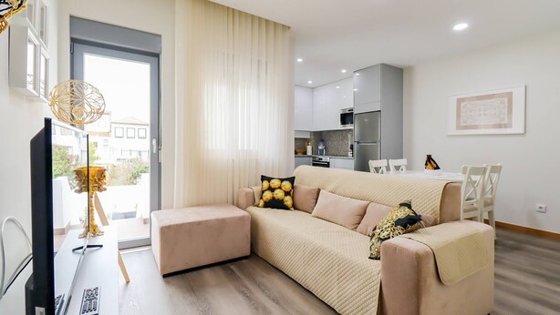 A modern living room with a beige sofa, a flat-screen TV, and a dining area in the background.