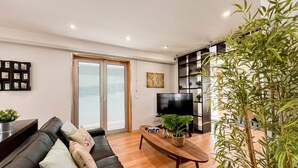Ein modernes Wohnzimmer mit einem schwarzen Ledersofa, einem Holztisch, einem Fernseher und einem großen Fenster mit Jalousien.