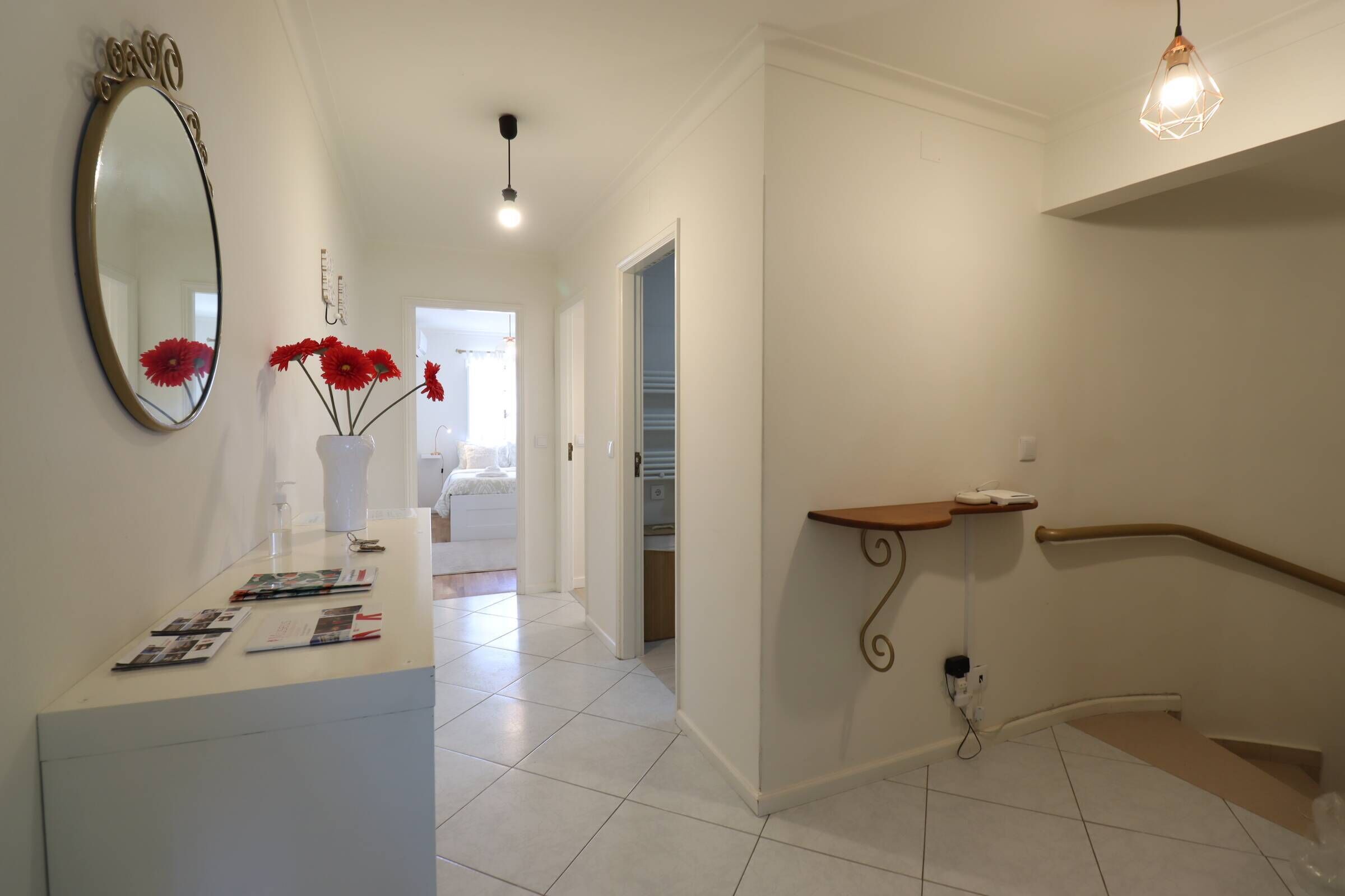 A hallway with a white tiled floor, a white reception desk with brochures, a mirror, and a vase with red flowers. There is a wooden bench with a metal frame and a light fixture above it.
