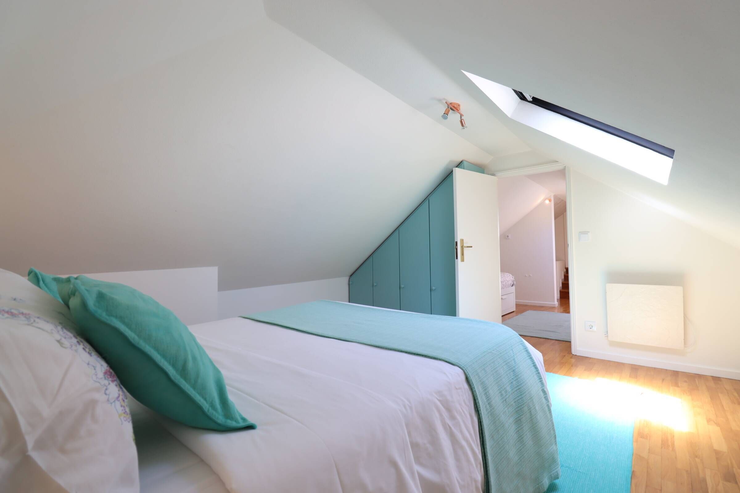 A bedroom with a bed, a skylight, a wardrobe, and a door leading to another room.