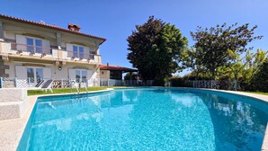 A clear blue swimming pool with a stone border, surrounded by a white fence and a building with balconies.