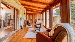 Une pièce chaleureuse avec un plancher en bois, une cheminée, un canapé et de grandes fenêtres donnant sur l’extérieur.