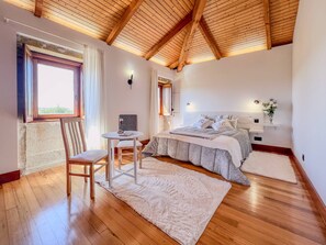 Ein Schlafzimmer mit Holzboden, einem Bett mit grauer Bettdecke, einem Nachttisch, einem Stuhl, einem kleinen Tisch und einem Fenster mit Vorhängen.