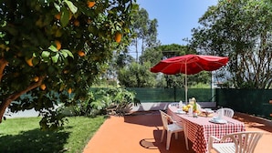Une terrasse extérieure avec une nappe à carreaux, des chaises blanches, un parasol rouge et une table dressée avec de la nourriture et des boissons.