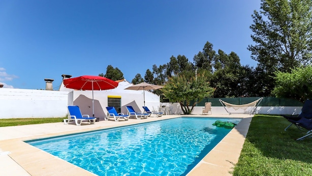Une piscine rectangulaire à l’eau d’un bleu limpide, entourée d’une terrasse carrelée. On y trouve des chaises longues bleues et un parasol rouge qui offre de l’ombre.