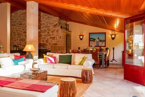 A cozy living room with a stone wall, a white sofa, a wooden ceiling, and a dining area in the background. - Justininho House - Luxurious Retreat (Ponte de Lima)