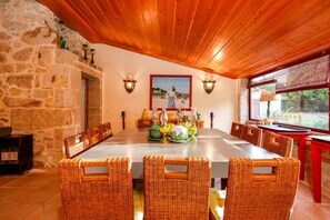 A dining area with a stone wall, wooden ceiling, wicker chairs, a long table, and wall-mounted lights. - Justininho House - Luxurious Retreat (Ponte de Lima)