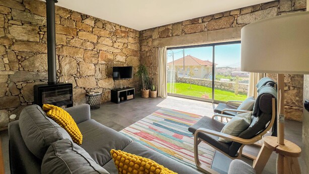 Un salon moderne avec un mur en pierre, une cheminée, un canapé avec des coussins jaunes, un téléviseur et une vue sur une maison à travers de grandes fenêtres.