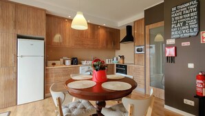 A modern kitchen with wooden cabinets, a central island with a red vase, and a dining area with a round table and cushioned chairs. - Da Silva House - Ap in Afife with Balcony & SeaView (Viana do Castelo)